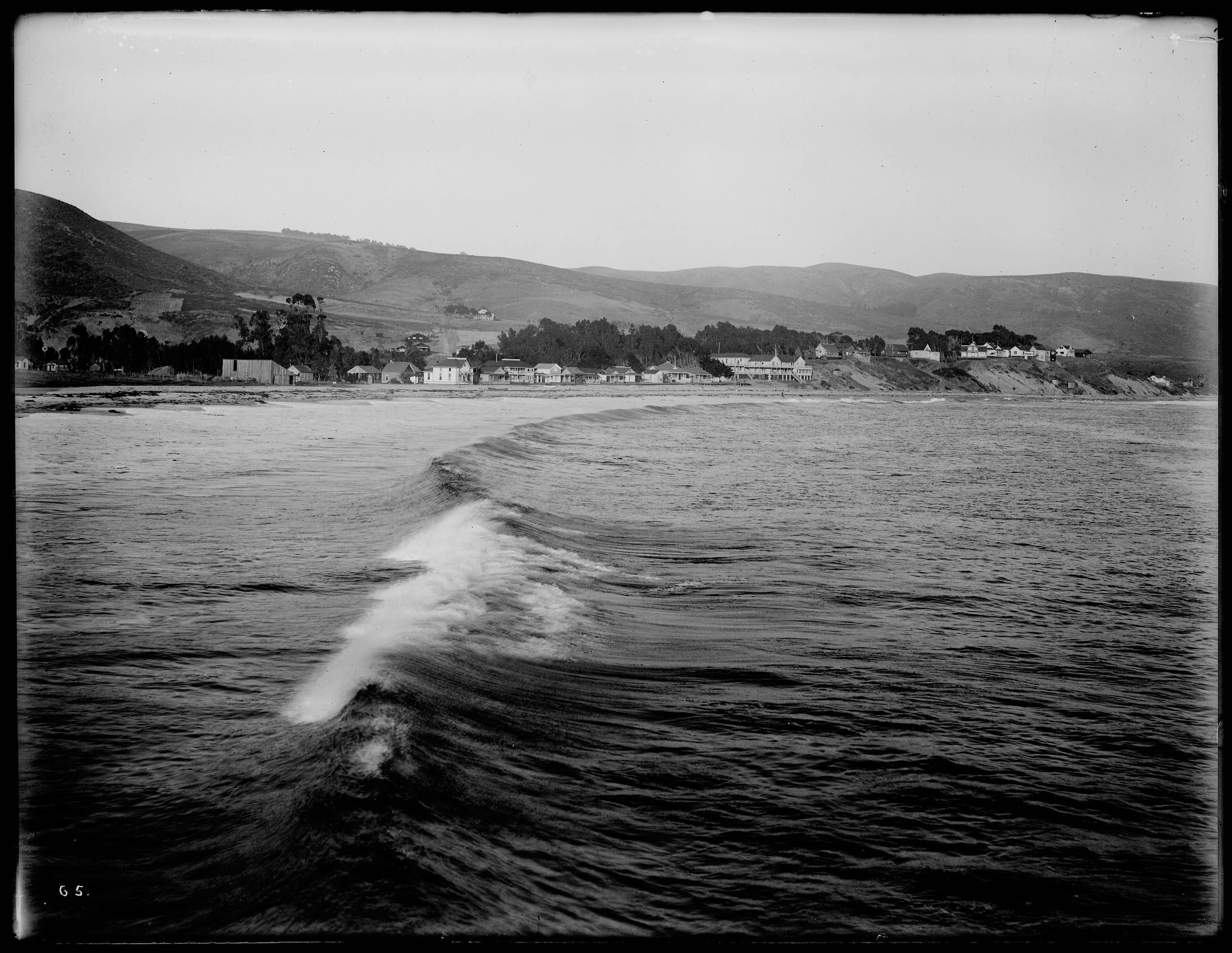 Black-and-white photograph of a breaking wave off Laguna Beach with small early houses along the shore and bare hills behind, about 1909