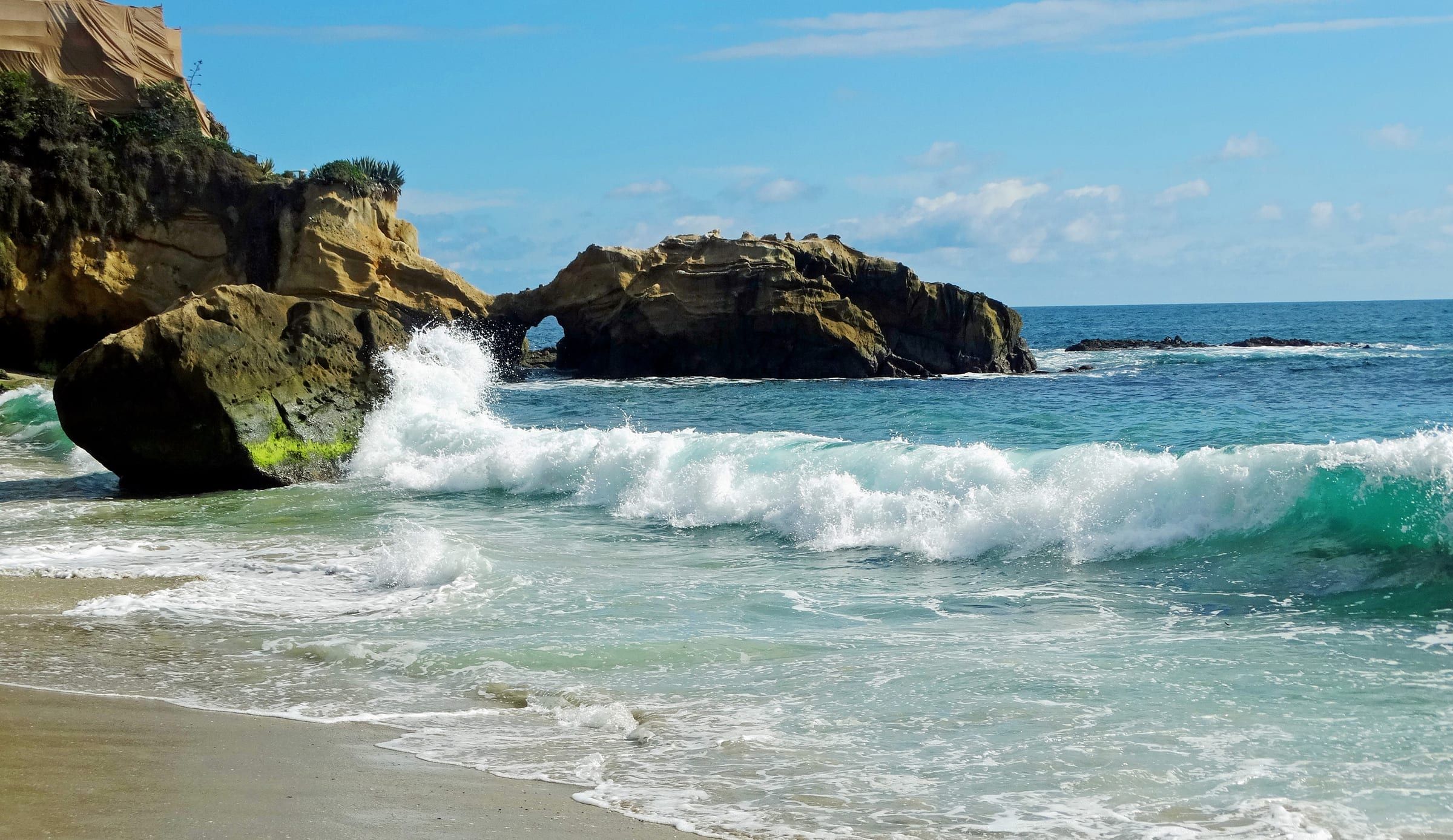 A wave breaking on a Laguna Beach shore, with a sea arch in an offshore rock behind it