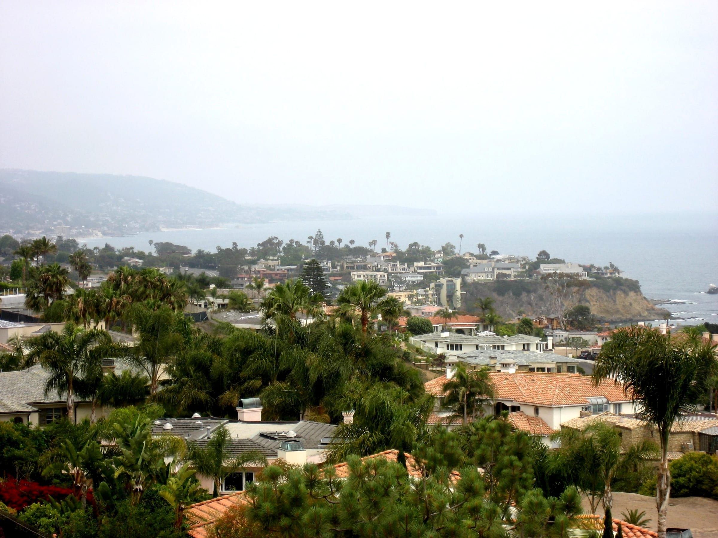 Laguna Beach seen from Crystal Cove on a hazy day, with hillside rooftops and palms in front and a headland and the ocean beyond