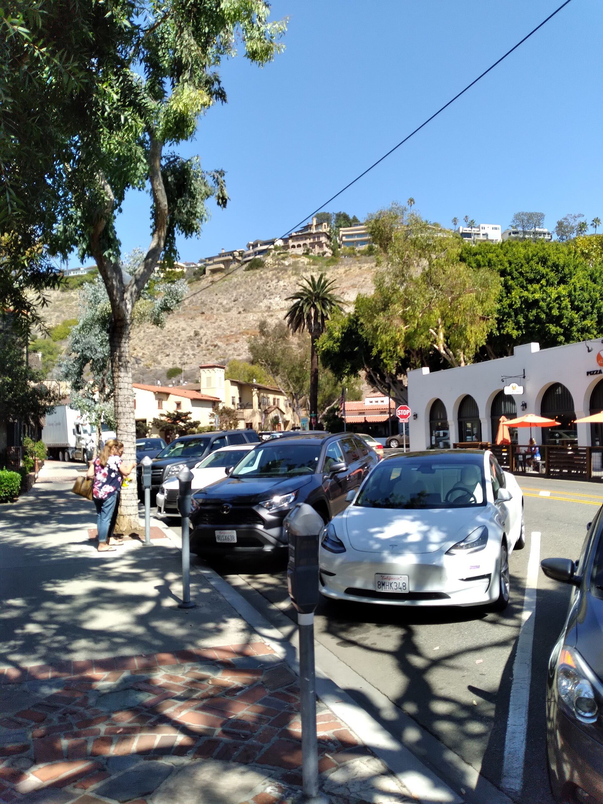 A downtown Laguna Beach street with parking meters, a brick sidewalk, a white arcaded building with patio umbrellas and hillside homes above