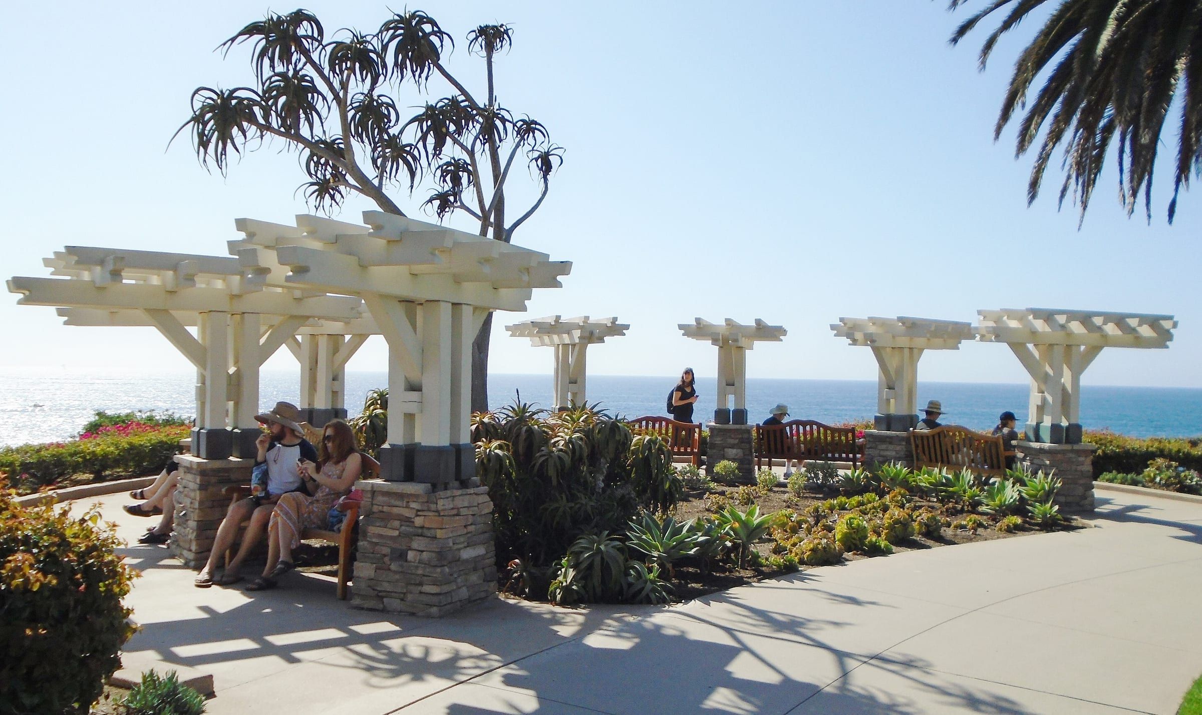 Visitors on benches under white pergolas along the Laguna Beach shoreline walkway, with the Pacific behind