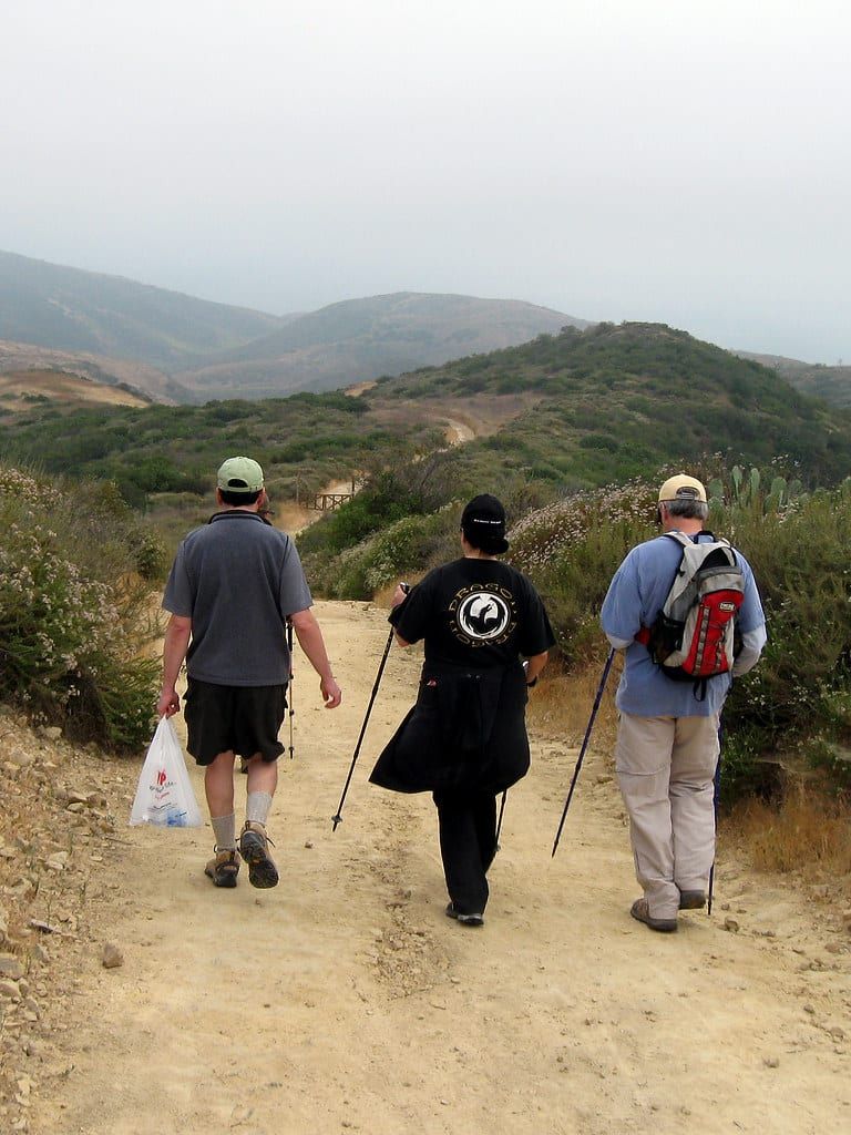 Three hikers, two with trekking poles, on a dirt trail through coastal hills in Crystal Cove State Park