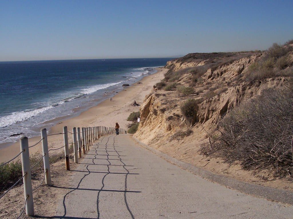 A paved ramp with a rope railing runs down a sandy bluff to a long beach with surf at Crystal Cove State Park